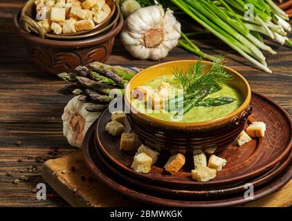 Cremige Spargelsuppe in Glasschale auf schwarzem Teller vor rohem frischem Spargel und Grün auf rustikalem Holzhintergrund Stockfoto