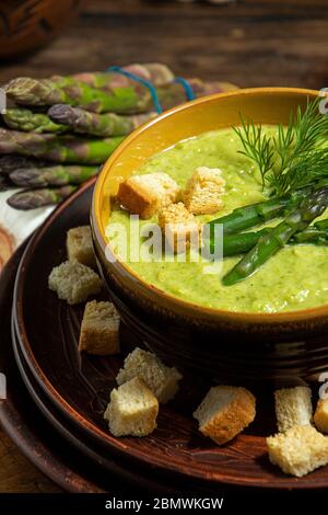 Cremige Spargelsuppe in Glasschale auf schwarzem Teller vor rohem frischem Spargel und Grün auf rustikalem Holzhintergrund Stockfoto