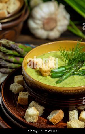 Cremige Spargelsuppe in Glasschale auf schwarzem Teller vor rohem frischem Spargel und Grün auf rustikalem Holzhintergrund Stockfoto