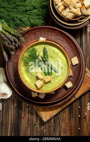 Cremige Spargelsuppe in Glasschale auf schwarzem Teller vor rohem frischen Spargel und Grün auf rustikalem Holzhintergrund. Draufsicht Stockfoto