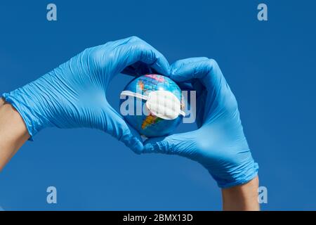 Mann, mit blauen OP-Handschuhe, die ein Herz mit seinen Händen, hält eine terrestrische Kugel mit einer Schutzmaske, die Darstellung der globalen Coronaviru Stockfoto