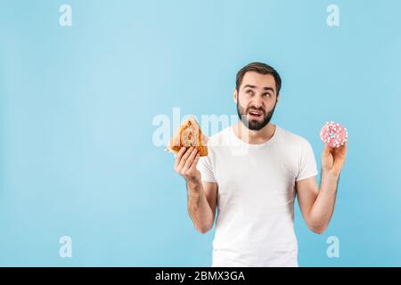 Bild von jungen denken verwirrt Mann isoliert über blaue Wand Hintergrund hält Sandwich und Donut. Stockfoto