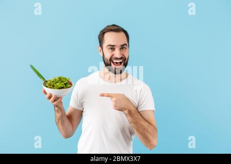 Schöner fröhlicher junger bärtiger Mann, der T-Shirt trägt, isoliert auf blauem Hintergrund stehend, Schüssel mit frischem Salat haltend, Zeigefinger Stockfoto