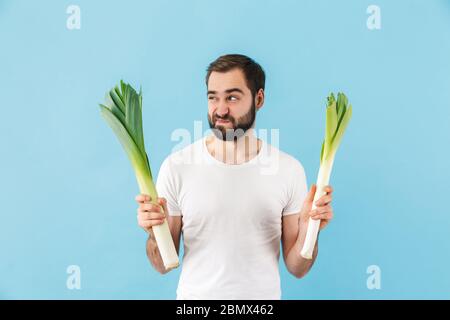 Schöner, verärgert junger bärtiger Mann, der T-Shirt trägt, isoliert auf blauem Hintergrund stehend, Lauch zeigt Stockfoto