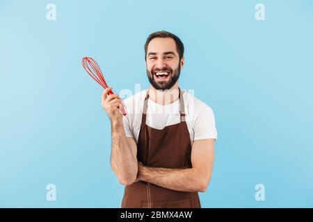 Fröhlicher junger bärtiger Mann mit Schürze, der isoliert auf blauem Hintergrund steht Stockfoto