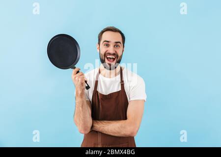 Aufgeregt jungen bärtigen Mann Koch trägt Schürze stehend isoliert über blauem Hintergrund, zeigt Bratpfanne Stockfoto