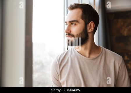 Schöner, ernster junger bärtiger Mann, der zu Hause am Fenster steht und wegschaut Stockfoto