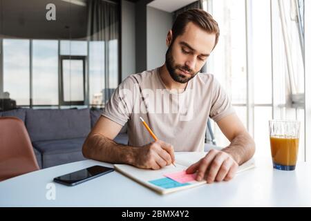Bild eines konzentrierten ernsten jungen Mannes zuhause zu Hause Schreiben Notizen sitzen am Tisch. Stockfoto