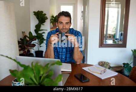 Lächelnder junger Mann, der Kaffee trinkt, während er von zu Hause aus arbeitet Stockfoto