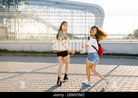 Verrückte lustige Foto von zwei fröhlichen asiatischen jungen Frauen in stilvollen Sommerkleidung, Laufen und Spaß im Freien in der Nähe des modernen Gebäudes mit großem Glas Stockfoto