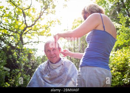 Die isolierende Schwiegertochter gibt ihrem Schwiegervater während der Sperrung im Garten während der Coronavirus-Pandemie einen Haarschnitt. Stockfoto