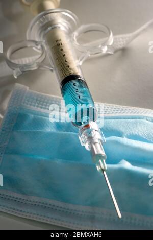 Konzeptbild einer Spritze mit blauer Flüssigkeit auf einer blauen PSA-Gesichtsmaske. Könnte zur Veranschaulichung der COVID-19-Pandemie verwendet werden. Stockfoto