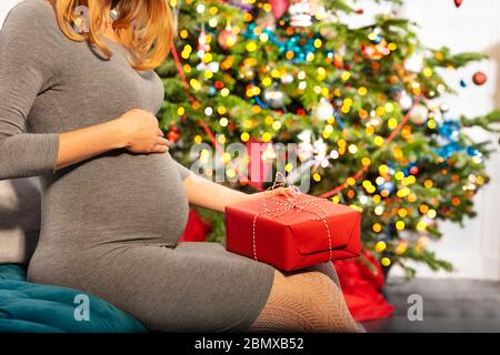 Nahaufnahme des Bauches der schwangeren Frau, die mit Geschenken auf dem Sofa in der Nähe des Neujahrsweihnachtsbaums sitzt Stockfoto