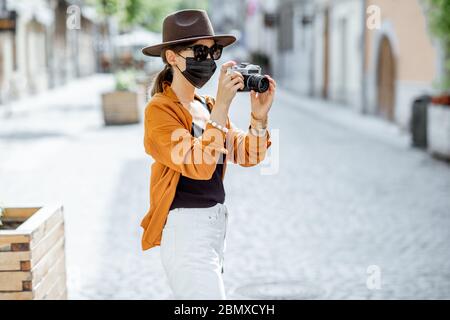 Junge weibliche Touristen in Gesichtsschutzmaske zu Fuß mit Fotokamera auf der alten Stadtstraße. Konzept des Tourismus und neue soziale Regeln und Einschränkungen nach Coronavirus-Pandemie Stockfoto