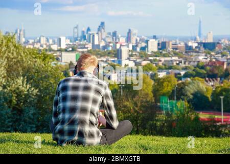 Rückansicht eines Mannes, der auf Gras sitzt und die Skyline von London vom Parliament Hill in Hampstead Heath aus betrachtet Stockfoto