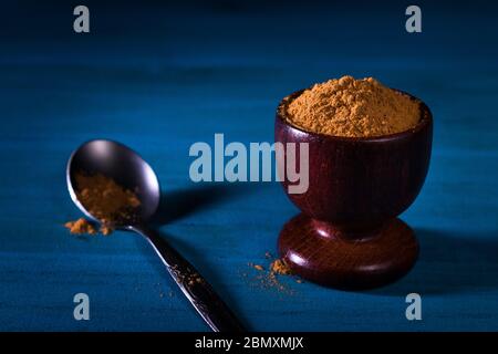 Gemahlenes Zimtpulver in Holzbecher mit kleinem Metalllöffel auf blauem Holztisch mit dunklem Hintergrund. Nahaufnahme. Dieses Bild oder ähnliches hat man nicht Biene Stockfoto