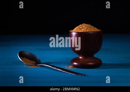 Gemahlenes Zimtpulver in Holzbecher mit kleinem Metalllöffel auf blauem Holztisch mit dunklem Hintergrund. Nahaufnahme. Stockfoto