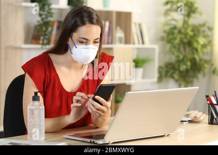 Mädchen mit Laptop, das Smartphone in Covid-19-Haft überprüft, und zu Hause auf einem Schreibtisch sitzt Stockfoto