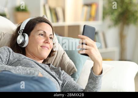 Glückliche Erwachsene Dame mit Kopfhörer hört Musik auf Smartphone liegen auf einer Couch zu Hause Stockfoto