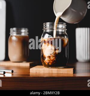 Herstellung von Eislaten. Vegetarische Sojamilch in ein Glas mit schwarzem Kaffee und Eiswürfeln gießen. Stockfoto