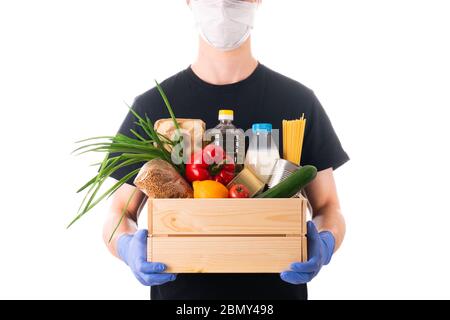 Junger Mann in einer Maske und einem schwarzen T-Shirt und Latexhandschuhe hält eine Holzkiste mit Lebensmitteln für die Lieferung isoliert auf weißem Hintergrund. Spendenbox Stockfoto