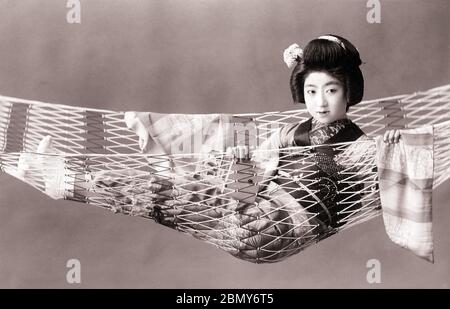 [ Japan der 1920er Jahre - Maiko in Hängematte ] - EIN Maiko (Lehrling Geisha) in Kimono posiert in einer Hängematte. Sie hält ein kleines Buch. Diese Postkarte wurde vom Kamigataya Studio in Tokio zwischen März 1918 (Taisho 7) und Februar 1933 (Showa 8) veröffentlicht. Vintage-Postkarte des 20. Jahrhunderts. Stockfoto