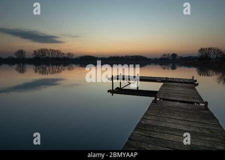 Hölzerner Angelsteg an einem ruhigen See, Himmel nach Sonnenuntergang und eine kleine Wolke Stockfoto