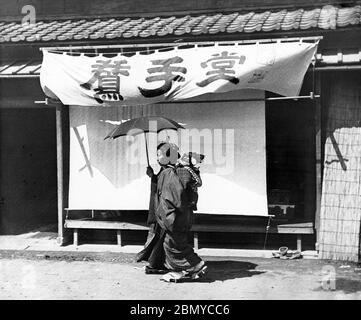 [ 1890er Jahre Japan - Japanische Mutter und Kind ] - Mutter mit einem Kind auf dem Rücken (onbu) vor einem Geschäft. Aus einer Serie von Glasdias, die vom schottischen Fotografen George Washington Wilson (1823–1893) veröffentlicht (aber nicht fotografiert) wurden. Wilsons Firma war einer der größten Verleger von Fotodrucken in der Welt. 19. Jahrhundert Vintage Glas Rutsche. Stockfoto