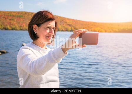 Frau, die an einem sonnigen Herbsttag ein Selfie auf einem Bergsee nimmt. Atemberaubende Herbstfarben im Hintergrund. Stockfoto