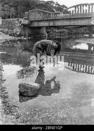 [ 1946 Japan - Okinawan Frau, die ihr Haar wäscht ] - Junge Okinawan Frau, die in einer Hose ihre Haare in einem Fluss in Taira (田井等) in Nago (名護), Okinawa, 1946 (Showa 21) trägt. Die Brücke hinter ihr trägt noch die Narben des Krieges. Silberdruck mit Gelatine aus dem 20. Jahrhundert. Stockfoto