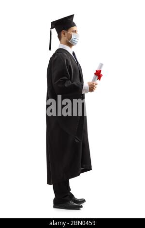 In voller Länge Profil Aufnahme eines männlichen Studenten in einem Abschlusskleid trägt eine schützende Gesichtsmaske und hält ein Diplom isoliert auf weißem Hintergrund Stockfoto