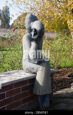 Eine Skulptur einer sitzenden jungen Frau, die ein Buch im Rosengarten in der Nähe der Tercentennary Sundial, Herstmonceux Castle Gardens liest Stockfoto