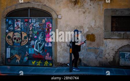 Eine nicht identifizierbare Dame mittleren Alters in Laufschuhen verwendet ein Mobiltelefon in einer rustikalen italienischen Straße, die an einem Bogengang vorbeiführt Der Eingang ist mit Straßenkunst überzogen Stockfoto