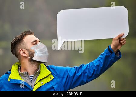 Ein bärtiger junger Mann, der eine medizinische Schutzmaske trägt, hält ein leeres Pappblatt auf einem verschwommenen natürlichen Hintergrund in der Hand. Ein weißes Männchen trägt ein Stockfoto