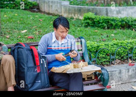 Ein junger Mann sitzt auf einer Parkbank, die Bambus schneidet und touristische Souvenirs in der Nähe des Hoan Kiem Sees, der Innenstadt von Hanoi, Nord-Vietnam, Südostasien macht Stockfoto