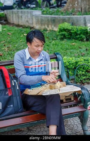 Ein junger Mann sitzt auf einer Parkbank, die Bambus schneidet und touristische Souvenirs in der Nähe des Hoan Kiem Sees, der Innenstadt von Hanoi, Nord-Vietnam, Südostasien macht Stockfoto