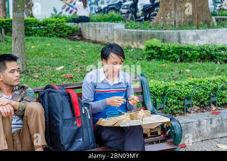 Ein junger Mann sitzt auf einer Parkbank, die Bambus schneidet und touristische Souvenirs in der Nähe des Hoan Kiem Sees, der Innenstadt von Hanoi, Nord-Vietnam, Südostasien macht Stockfoto