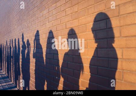 Die Menschen drängen Schatten aufgereiht gegen eine rote Ziegelmauer. Sie stehen in einer Schlange für Veränderungen im Leben. Soziale Distanz, Covid und Immigration Themenkonzept Stockfoto