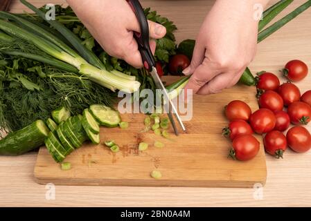 Frau schneidet grüne Zwiebel mit einer Schere über einem Schneidebrett. Grüns, Tomaten und Gurken auf einem Holztisch. Rustikaler Stil. Stockfoto