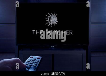 Ein Mann zeigt eine Fernbedienung auf den Fernseher, der den Hauptbildschirm des The Witcher-Titels anzeigt (nur redaktionelle Verwendung). Stockfoto