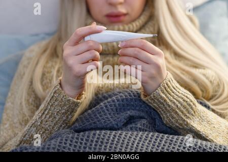 Kranke junge Frau mit Fieber liegt im Bett unter der Decke, prüft die Temperatur und schaut auf Thermometer, selektive Fokussierung. Erkältung, Virus, Grippeasuso Stockfoto
