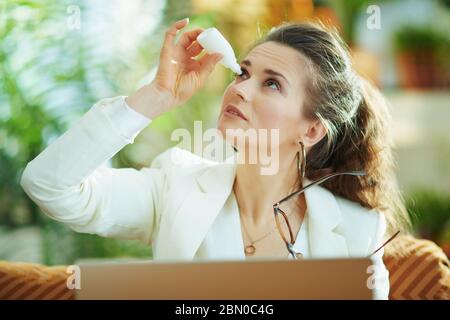 Moderne Hausfrau in weißer Bluse und Jacke mit Laptop und Brille mit Augentropfen im modernen Zuhause an sonnigen Tag. Stockfoto