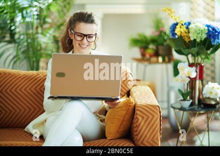 Glückliche junge Frau in weißer Bluse und Jacke in modernem Zuhause in sonnigen Tag Überprüfung Website auf einem Laptop während auf Diwan sitzen. Stockfoto