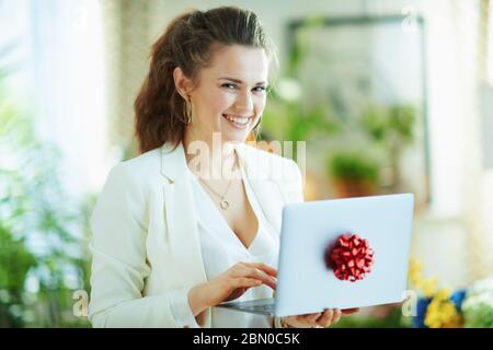 Portrait der lächelnden trendigen 40 Jahre alten Hausfrau in weißer Bluse und Jacke mit Geschenk-Laptop mit roter Schleife in der modernen Wohnzimmer bei schönem Tag. Stockfoto