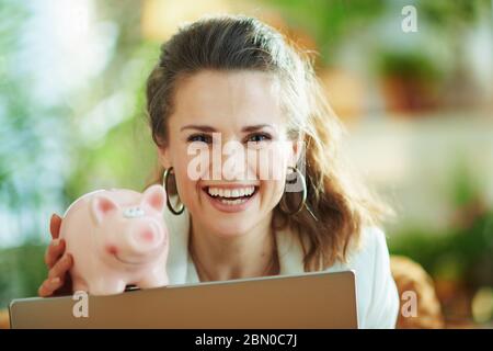 Portrait der lächelnden modernen 40 Jahre alten Frau in weißer Bluse und Jacke mit Laptop und Sparschwein im modernen Wohnzimmer bei schönem Tag. Stockfoto