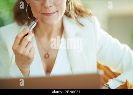 Nahaufnahme auf Frau in weißer Bluse und Jacke mit Laptop Anwendung Lippenstift während auf der Couch im Wohnzimmer an sonnigen Tag sitzen. Stockfoto