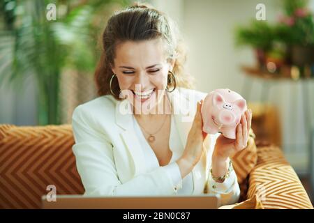 Lächelnde trendige Frau in weißer Bluse und Jacke mit Laptop und Sparschwein im modernen Wohnzimmer bei schönem Tag. Stockfoto