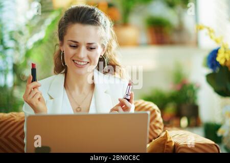 Happy elegante Frau in weißer Bluse und Jacke mit Laptop und zwei Lippenstifte vlogging in das moderne Haus an sonnigen Tag. Stockfoto