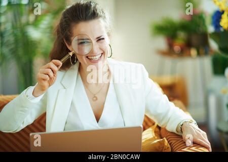 Portrait der fröhlichen trendigen Hausfrau in weißer Bluse und Jacke mit Laptop und Lupe bei modernem Zuhause an sonnigen Tag. Stockfoto