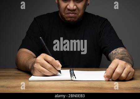 Asian Mann Schreiben auf einem Notizbuch mit wütenden Gesicht auf Holztisch. Konzept über Remote-Arbeit. Stockfoto